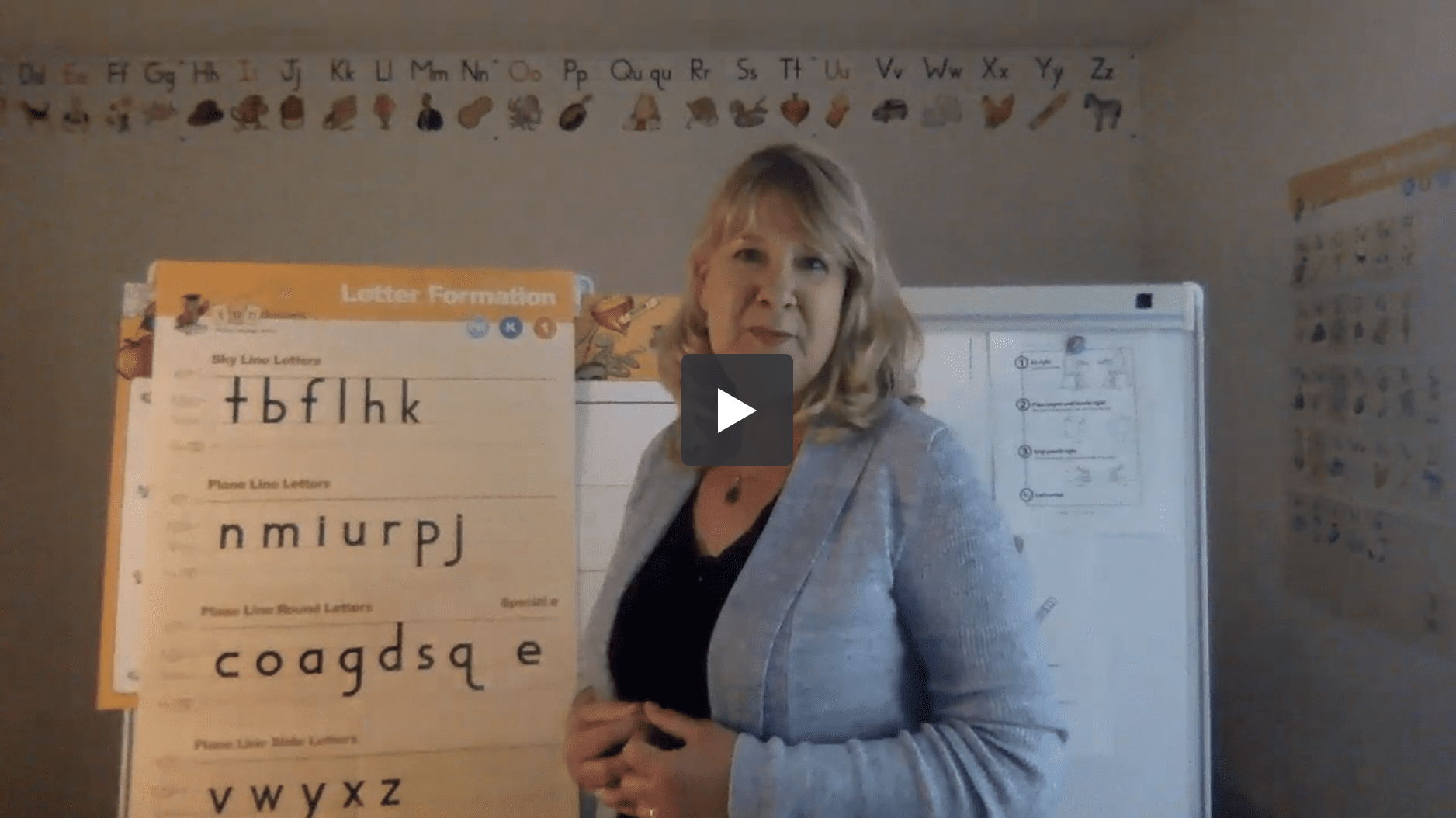 A woman is standing in a classroom with a large poster titled Letter Formation displaying letters. She wears a light blue blazer and smiles at the camera. The alphabet is displayed on the wall behind her. The play button in the center suggests it's a video.